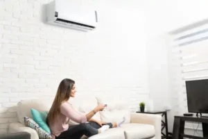 A woman sits on a sofa in a living room, holding a remote control and pointing it at a wall-mounted air conditioner.