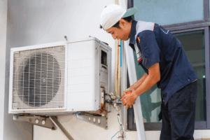 HVAC technician repairing an outdoor air conditioning condenser unit, adjusting electrical connections with tools beside a building window.