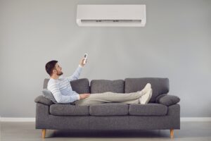 Man relaxing on a gray sofa while using a remote to control a wall-mounted air conditioner.