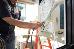 HVAC technician connecting refrigerant lines to an outdoor air conditioning unit during installation or maintenance, using tools and hoses beside a ladder.