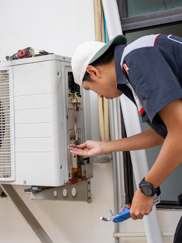 HVAC technician inspecting and servicing an outdoor air conditioning unit, checking internal wiring and refrigerant connections while holding a tool.